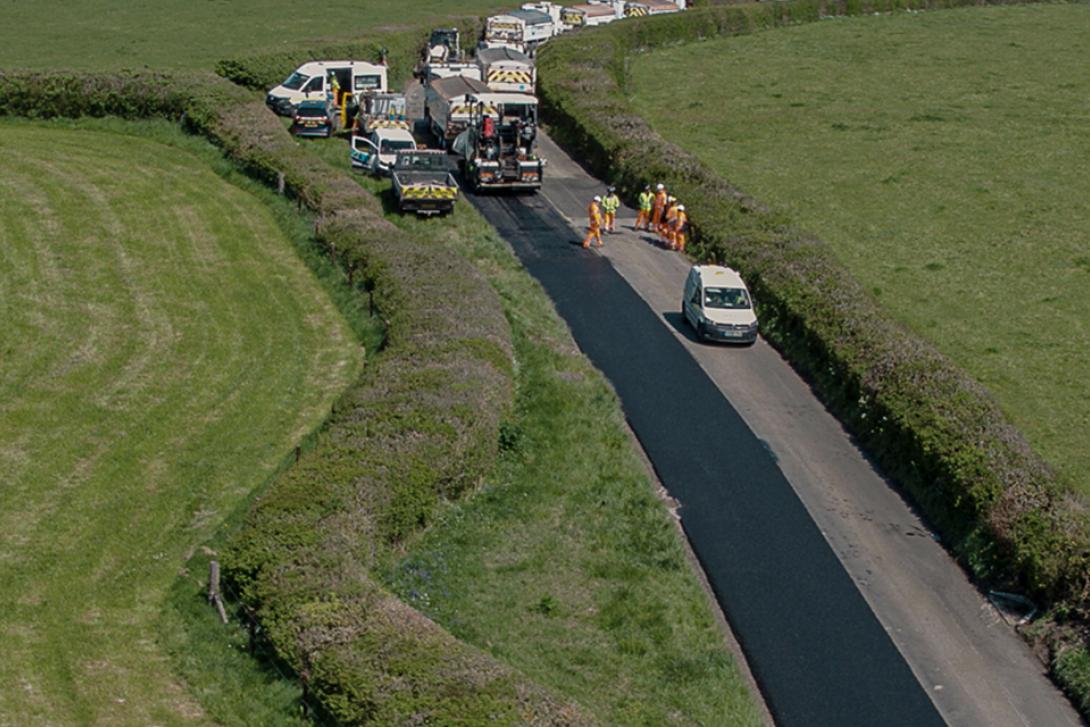 Road resurfacing in progress on a rural two-lane road surrounded by green hedges and fields; workers in high-visibility clothing operate machinery near freshly laid asphalt, while multiple utility vehicles are parked nearby.