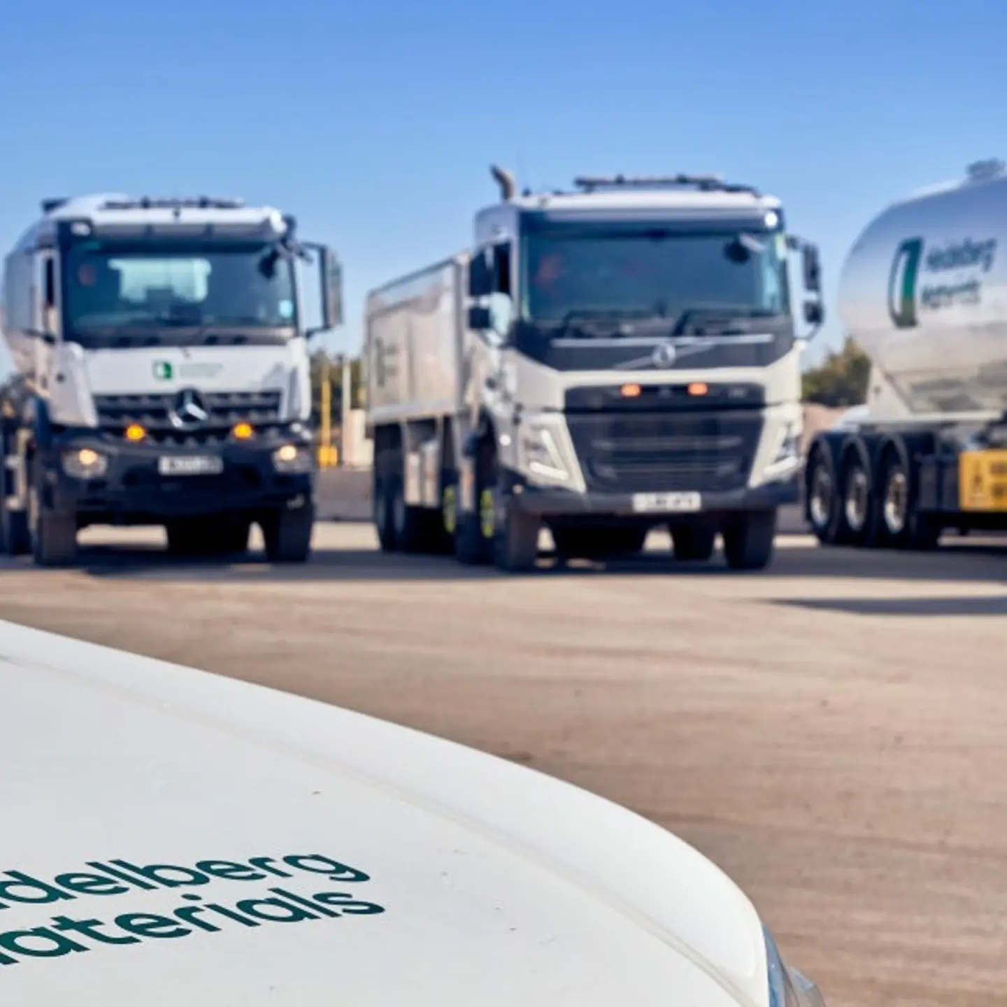 Four large commercial vehicles, including a cement tanker, concrete mixer are parked in a row on an open lot under a clear blue sky. 