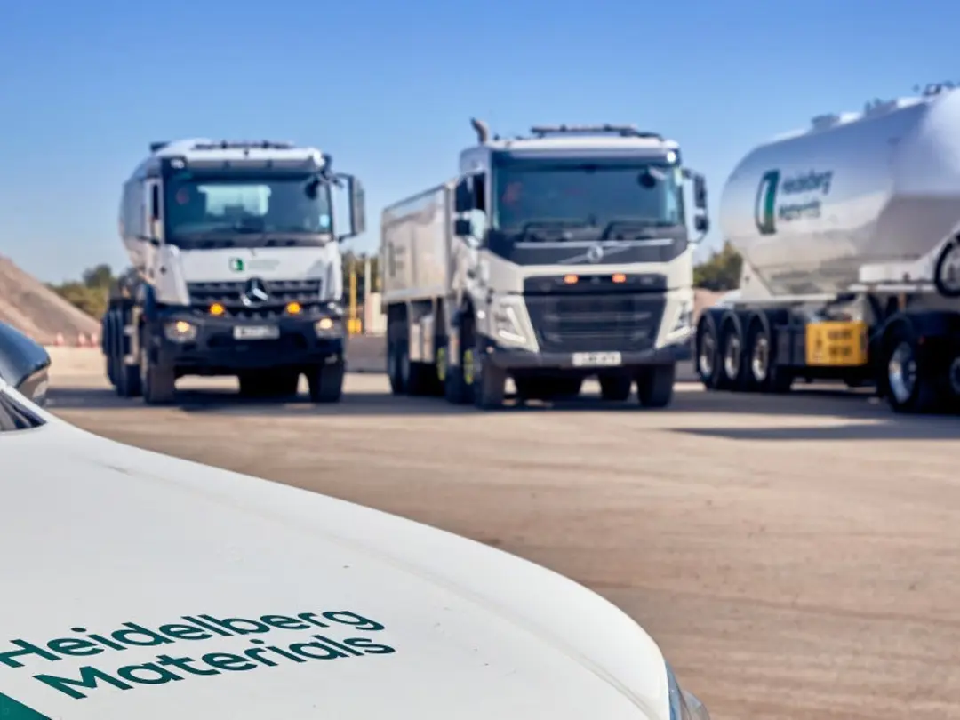 Four large commercial vehicles, including a cement tanker, concrete mixer are parked in a row on an open lot under a clear blue sky. 